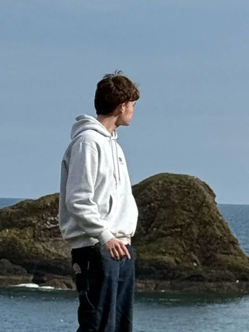A young man, Andrew Aslak Cruz Nimmo, standing on a grassy cliffside overlooking the North Sea with the ruins of Dunnottar Castle in the background.