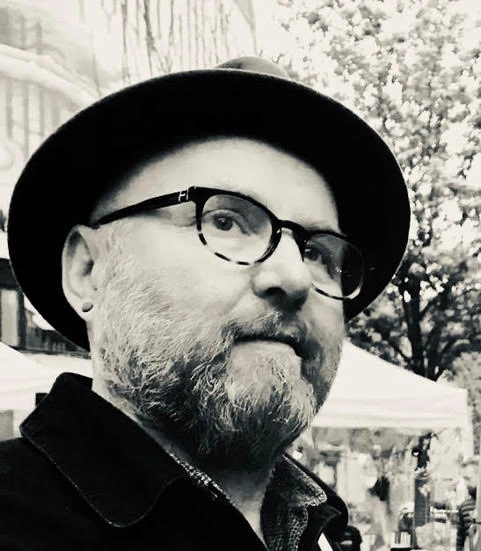 A black and white close-up portrait of Jim Ewen, an Aberdeen artist and musician, wearing a fedora hat and glasses.