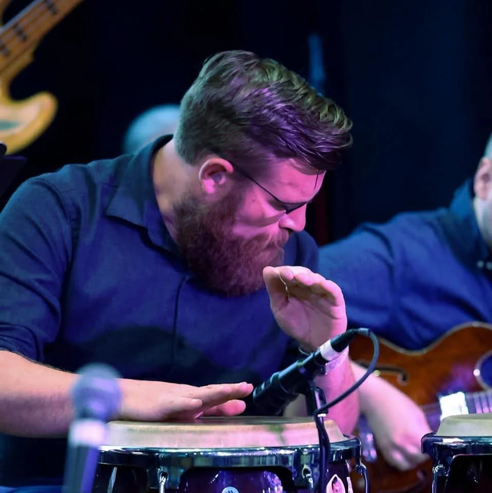 Michael Fenton, a bearded man wearing glasses and a navy shirt, playing the conga drums with focus during a live music performance.