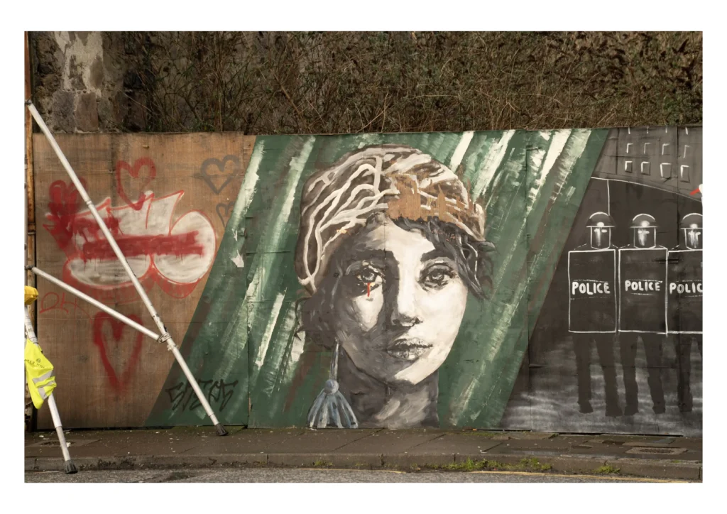 A large-scale street art mural on a wooden hoarding in Aberdeen, featuring a detailed monochrome portrait of a person with a single red tear, flanked by graffiti hearts and a stylised depiction of police officers.